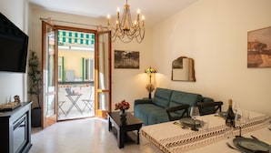 A living room with a sofa, dining table set for two, a chandelier, and a view through a glass door to an outdoor area.