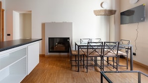 Une cuisine moderne dotée d’une cheminée intégrée, un coin repas avec une table en verre et des chaises en métal, et un téléviseur fixé au mur.