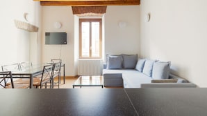 Un salon comprenant une table à manger en verre, un canapé, un téléviseur fixé au mur et une fenêtre avec des stores.