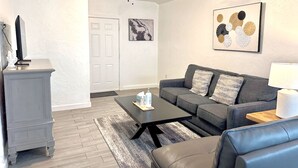 A living room with a gray sofa, a black coffee table, a fireplace, and a ceiling fan. - Fully-furnished Studio With Rustic Design 36B (Oklahoma City)