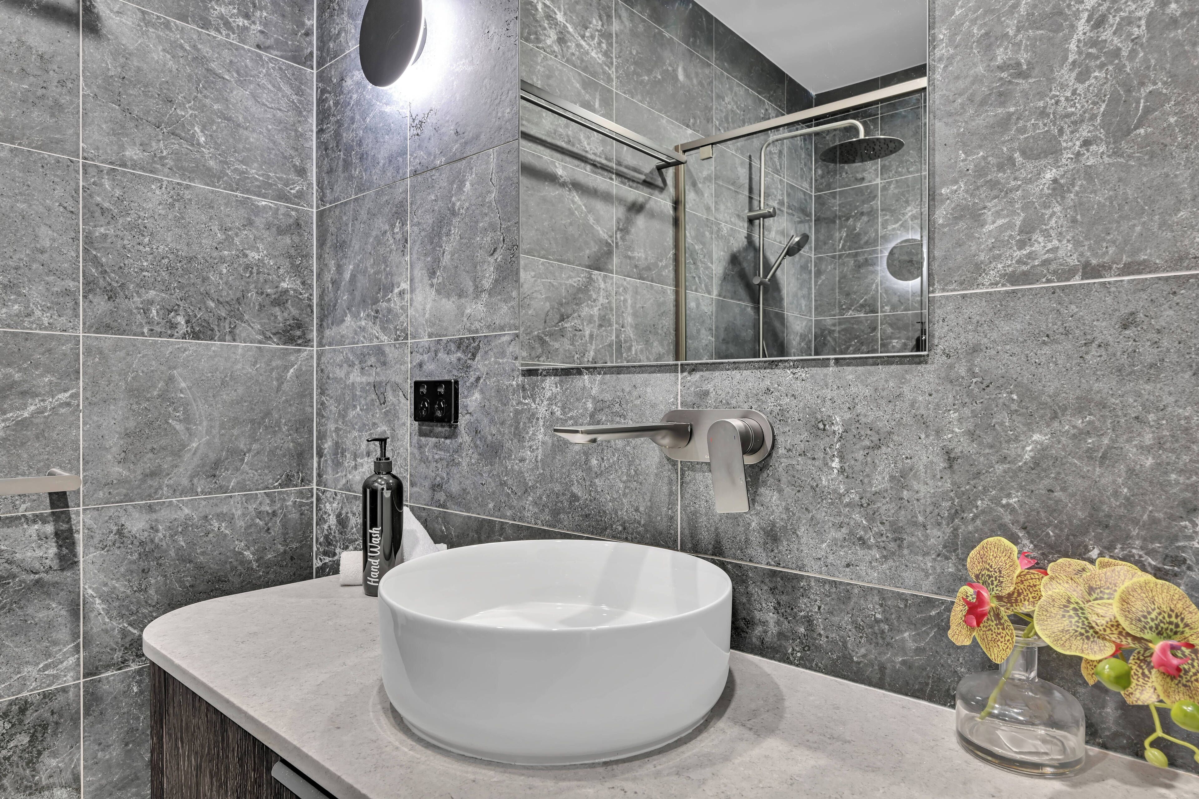Een moderne badkamer met een rond wit wastafel, een handdouche en een glazen vaas met orchideeën.