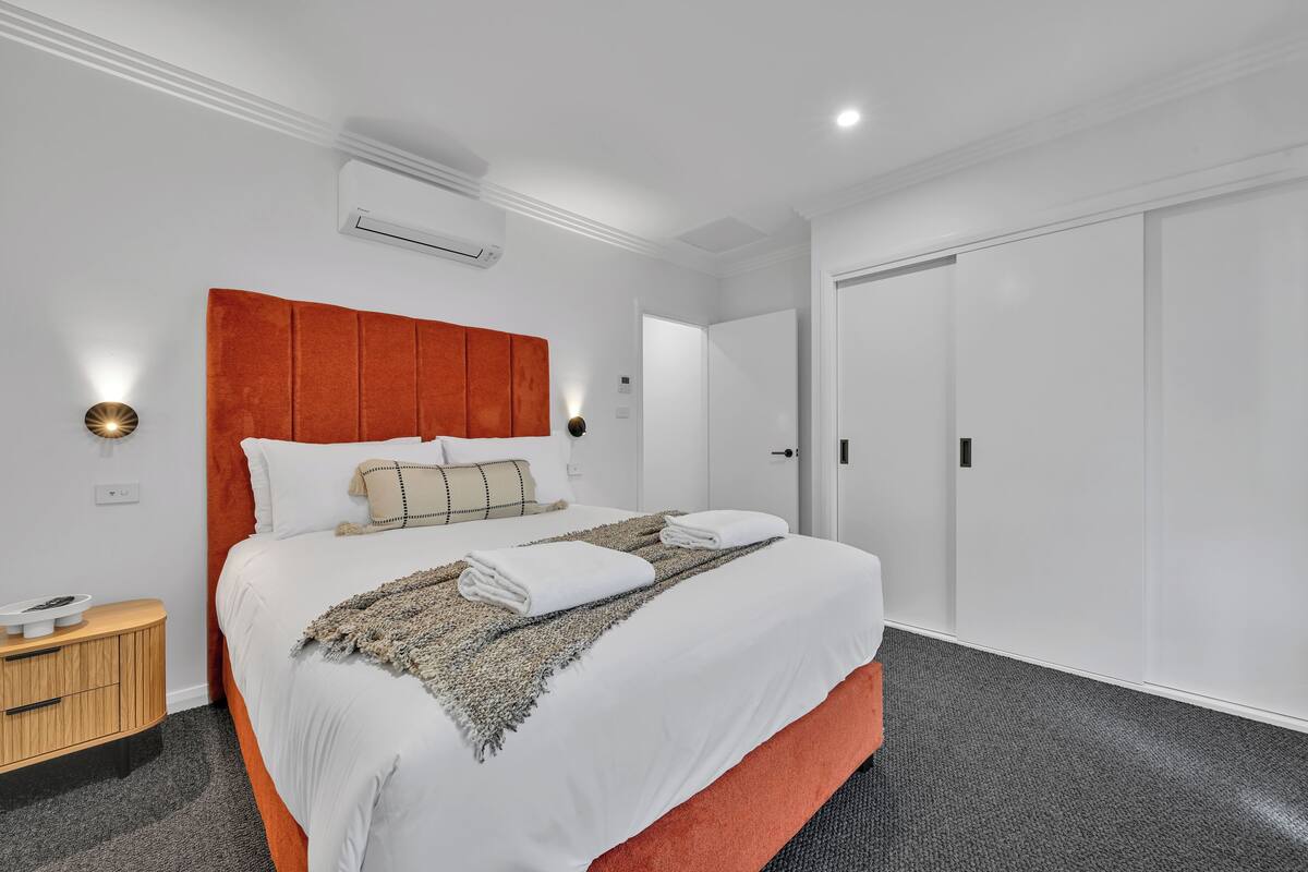 A neatly made bed with white linens and a red headboard, a bedside table with a lamp, and a grey carpeted floor.