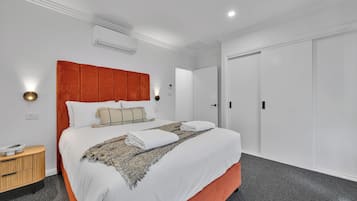 Una cama bien tendida con sábanas blancas y un cabecero rojo, una mesita de noche con lámpara y un suelo cubierto de moqueta gris.