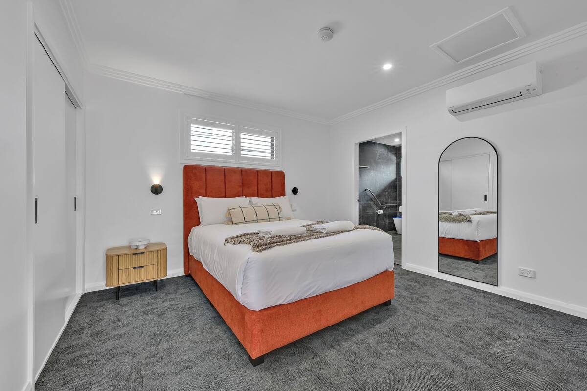 A bedroom with a bed, a nightstand, a mirror, and a window with blinds.