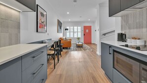 A modern kitchen with grey cabinets, a white countertop, and a built-in oven. There is a dining area with a sofa and chairs, and a red door in the background. - The Mac Orange (Orange)