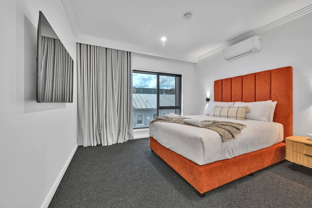 A modern bedroom with a large bed, a built-in TV, a window with curtains, and an air conditioning unit.
