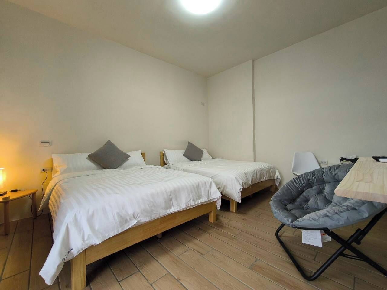 Una habitación con dos camas de madera, un sillón informal de tela gris y una mesita de noche con una lámpara.