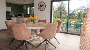 Ein moderner Essbereich mit einem runden Tisch und sechs Stühlen, im Hintergrund eine Küche und ein großes Fenster mit Blick ins Freie.