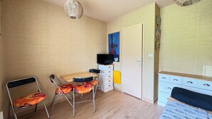 A room with a wooden dining table, two chairs, a television on a stand, a bed with patterned bedding, and a wall-mounted light fixture. - Appt Confortable in Superdévoluy (Le Dévoluy)
