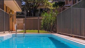 Piscina rectangular rodeada por una cerca metálica, con un edificio de ladrillo y una zona verde de fondo.