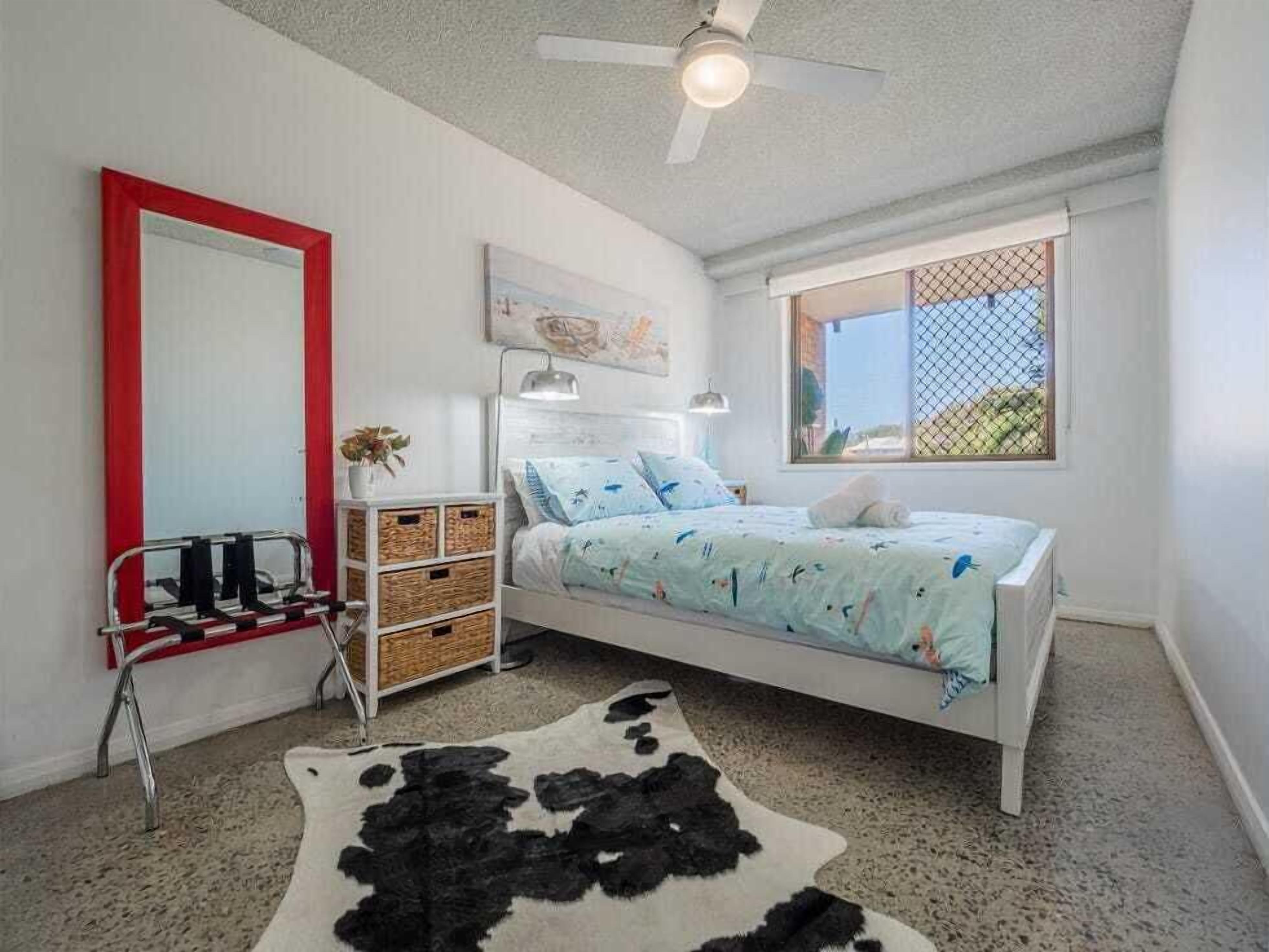 A bedroom with a bed, a mirror, a ceiling fan, a window, and a cowhide rug.