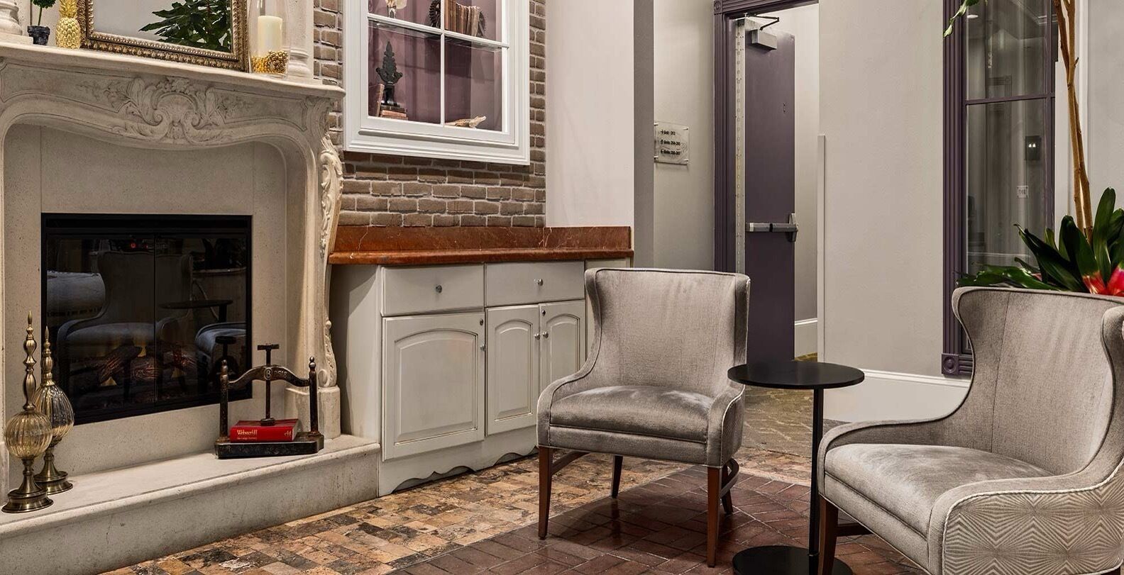 A cozy corner with a fireplace, two armchairs, a small table, and a window overlooking a brick wall.