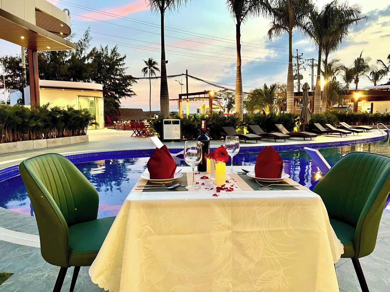 Mesa de comedor lista para dos personas, con vistas a la piscina, rodeada de sillones de descanso y palmeras.