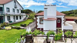 Un edificio blanco con puerta roja y tejado de tejas, rodeado por un césped cuidado y un pequeño jardín con parras.
