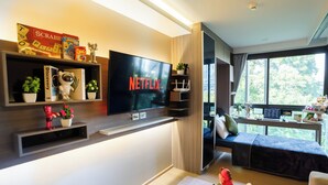 Una sala de estar moderna con un televisor de pantalla plana grande que muestra el logotipo de Netflix, una estantería con diversos objetos, una zona de estar acogedora con un sofá y una ventana con vistas a zonas verdes.