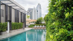 Una piscina moderna con sillas de descanso, rodeada de vegetación y edificios urbanos al fondo.