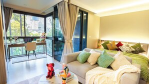 Una habitación de hotel moderna con balcón, un sofá con muchos cojines, una mesita de centro con un osito de peluche y un ventanal con cortinas.
