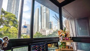 Un espacio de trabajo con dos monitores de ordenador que muestran gráficos, un escritorio con un ratón, y una ventana que ofrece vistas de edificios altos y árboles.
