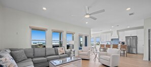 A spacious living area with a gray sofa, a coffee table, and a ceiling fan. There are large windows offering a view of the ocean.