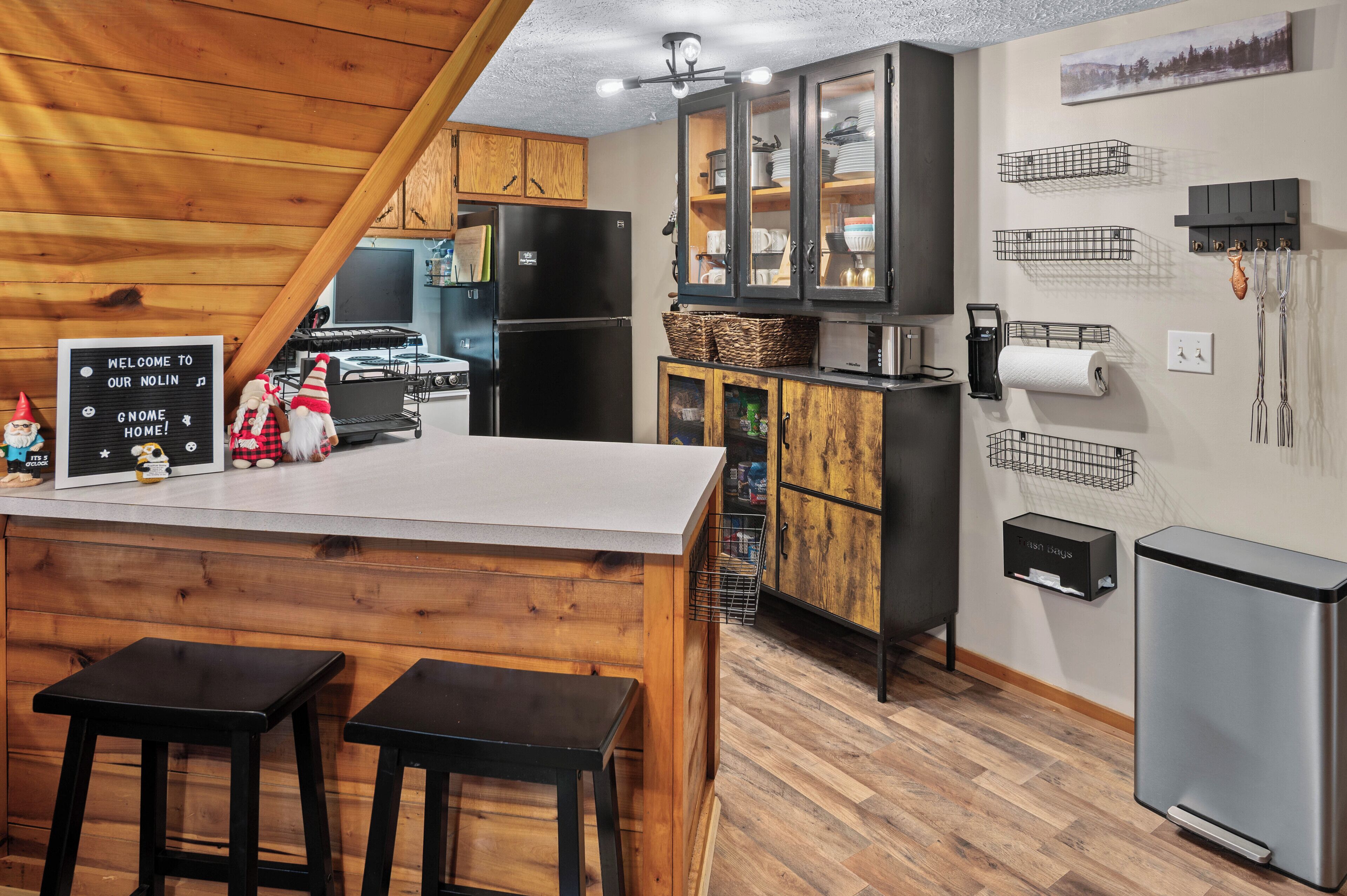 Una cocina con armarios de madera, un refrigerador negro, una isla de madera con taburetes y un cartel de bienvenida.