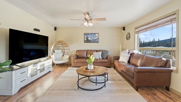 A living room with a flat-screen TV, two leather sofas, a round coffee table, a ceiling fan, and a large window with a view of trees and mountains.