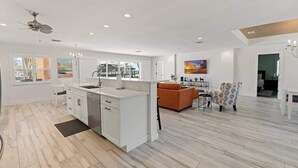 Une cuisine moderne avec des meubles blancs et un îlot central intégré, un coin repas avec une chaise à motifs et une grande fenêtre avec des stores.
