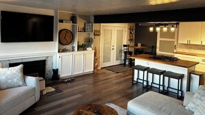 A modern living room with a fireplace, a TV mounted on the wall, and a kitchen area with a bar. - Charming 3-bedroom lake view home in tranquil Coeur d'Alene (Coeur d'Alene)