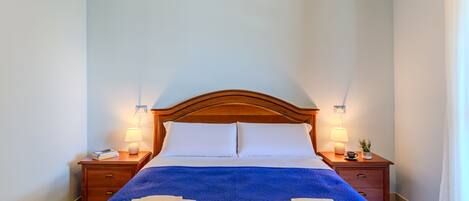 Una cama bien hecha con un edredón azul, dos almohadas blancas y dos toallas dobladas encima. Hay dos mesitas de noche, con lámparas y una pequeña maceta con planta en una de ellas. La habitación tiene paredes azul claro y un cabecero de madera.