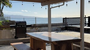 An outdoor patio with a wooden table and bench, a barbecue grill, string lights, and a view of the ocean. - Oceanfront Hideaway with Detached Primary Bedroom (Ewa Beach)