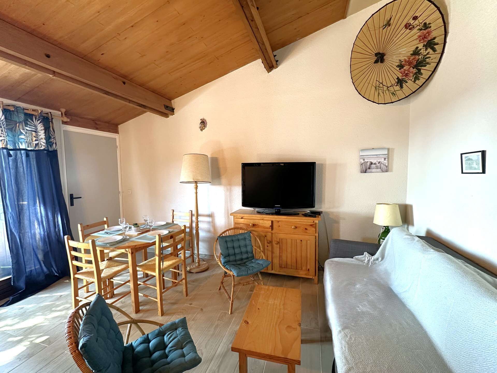 A cozy living room with a wooden ceiling, a dining area with wooden chairs and a table, a flat-screen TV on a wooden cabinet, and a large fan on the wall.
