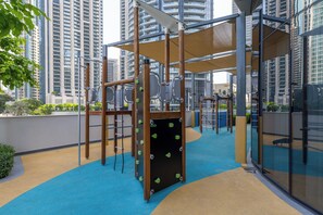 An outdoor play area with climbing structures and a blue and beige floor, surrounded by tall buildings. - Maison Privee Sky-High Elegance w/ Unrivaled Burj Khalifa Views (Dubai)