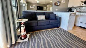 En moderne stue med en mørkeblå sofa, et lille rundt bord og en model af et fyrtårn. Et køkken med hvide skabe og en komfur kan ses i baggrunden.