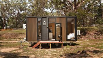 Una cabaña pequeña y moderna con entrada por puerta corrediza, un pequeño porche y una cocina visible con una parrilla.