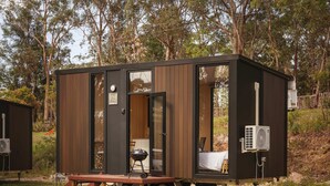 Una cabaña moderna y compacta con puerta corredera, un pequeño porche con una parrilla y un equipo de aire acondicionado.