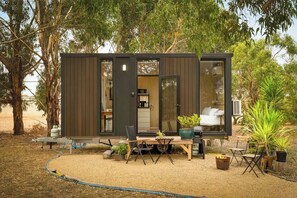 A modern, compact cabin with a dining area, kitchen, and sleeping quarters, surrounded by trees and outdoor furniture. - Bush Serenity by the Sea by Tiny Away (Bolwarra)