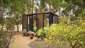 A modern, compact cabin with a wooden exterior and large windows, surrounded by a garden with various plants and trees.