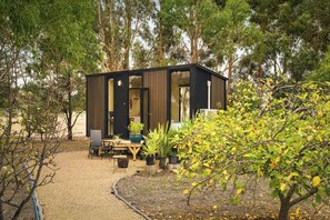 A modern, compact cabin with a wooden exterior and large windows, surrounded by a garden with various plants and trees. - Bush Serenity by the Sea by Tiny Away (Bolwarra)