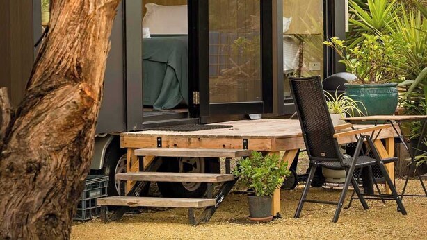 Una cabaña moderna y compacta con puerta corrediza de vidrio que da a un dormitorio. Cuenta con una pequeña terraza con mesa y sillas, rodeada de plantas en macetas.
