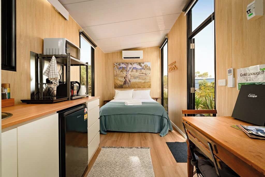 Ein kleines Hotelzimmer mit Küchenzeile, einem Bett mit grüner Bettdecke, einem Bild an der Wand und Blick durch das Fenster.