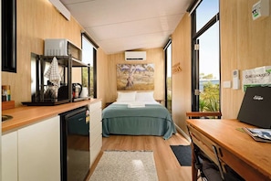 A compact hotel room with a kitchenette, a bed with a green bedspread, a painting on the wall, and a view through the window. - Bush Serenity by the Sea by Tiny Away (Bolwarra)