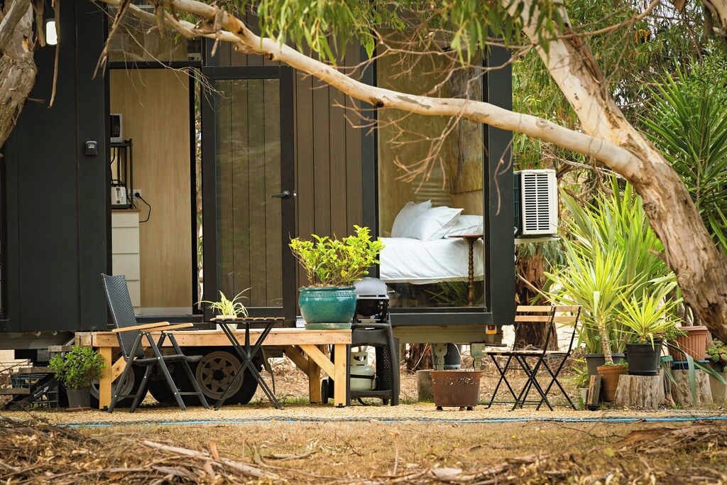 A small, enclosed outdoor cabin with a bed, a table, and chairs, surrounded by potted plants and a tree.