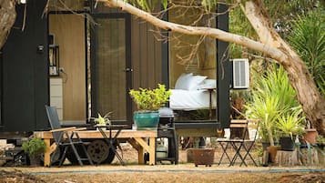 A small, enclosed outdoor cabin with a bed, a table, and chairs, surrounded by potted plants and a tree.