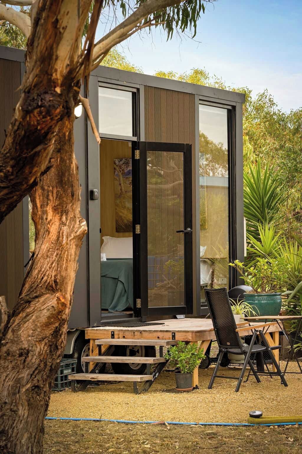 A modern, compact cabin with a sliding glass door leading to a bedroom. There is a small deck area with a table and chairs, surrounded by potted plants.