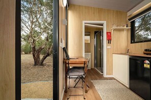 A compact kitchen with a wooden table and chairs, a black refrigerator, and a window overlooking a tree-lined outdoor area. - Bush Serenity by the Sea by Tiny Away (Bolwarra)