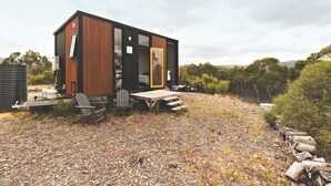 Una cabaña moderna y compacta con fachada de madera, zona de comedor con mobiliario exterior y vistas despejadas del paisaje circundante.