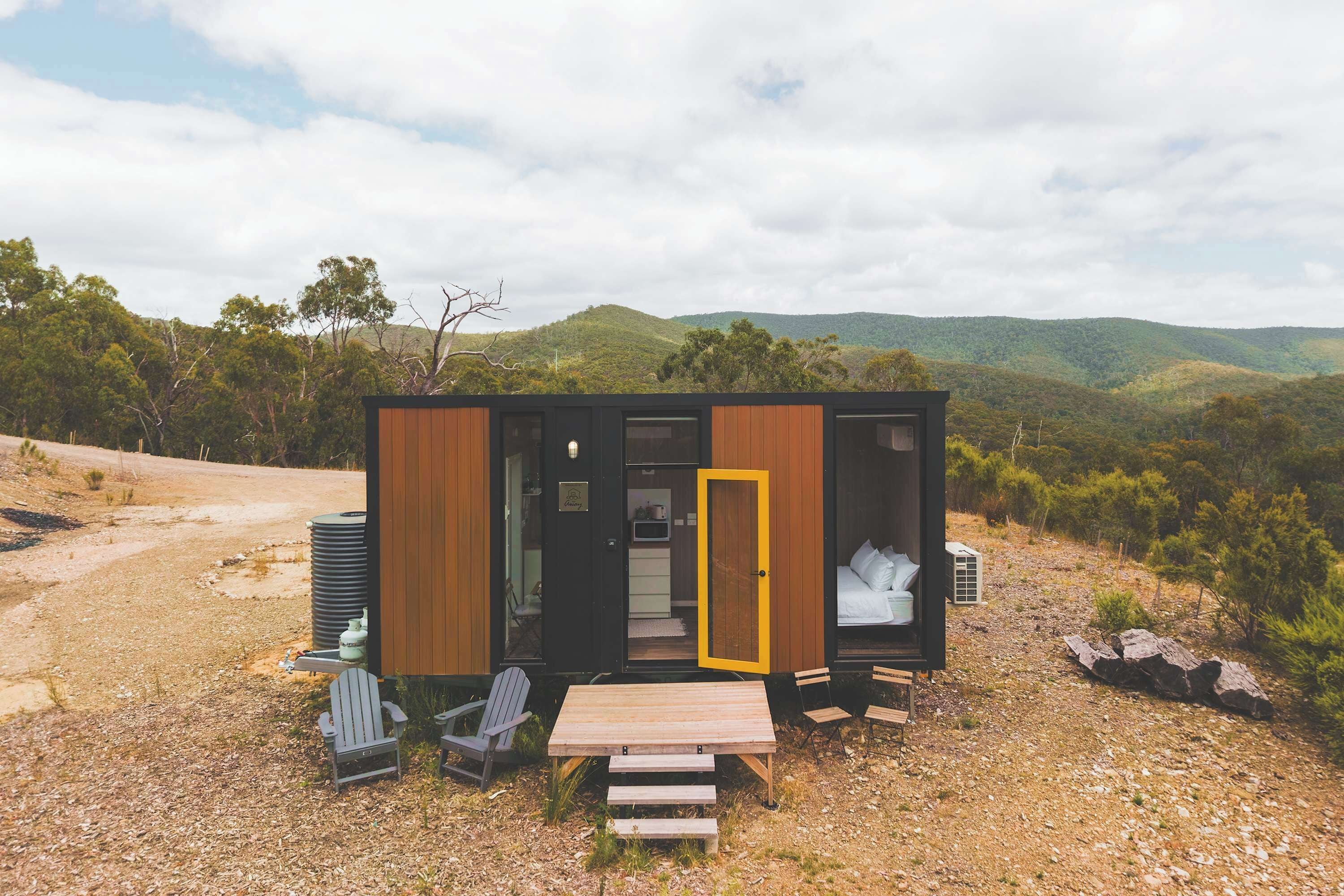 A compact cabin with a wooden exterior, a yellow door, and a small outdoor seating area with a table and chairs.