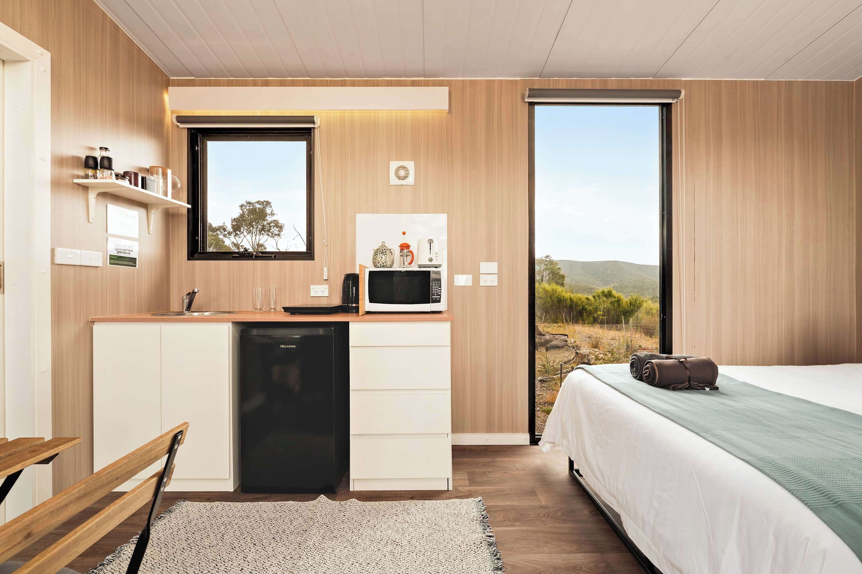 Ein modernes Schlafzimmer mit einem Bett, einem kleinen Schreibtisch mit Mikrowelle und Blick ins Freie.