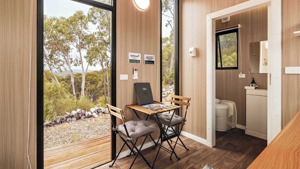 Una habitación moderna con interior de madera, una mesa de comedor con dos sillas, una pequeña cocina y un gran ventanal con vistas a árboles y una masa de agua.