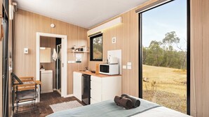 Un dormitorio moderno con cama, escritorio, una kitchenette pequeña y un amplio ventanal corredero al exterior.