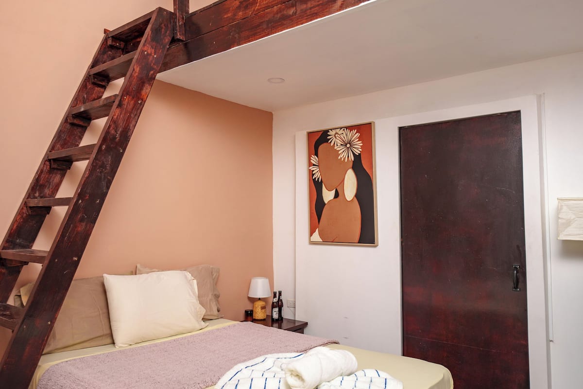 A bedroom with a bed, a nightstand, a lamp, a painting, and a wooden staircase.